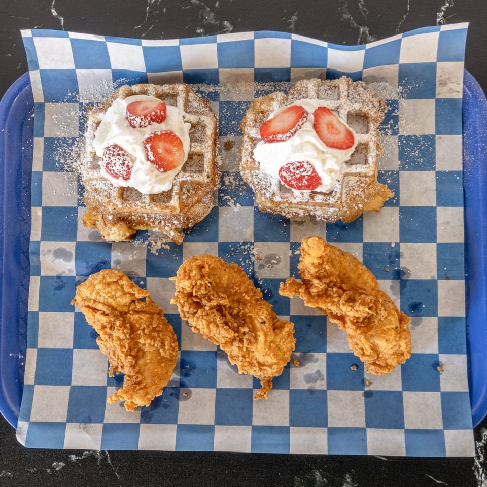 Two waffles with whipped cream and strawberries, and three pieces of fried chicken on blue checkered paper.