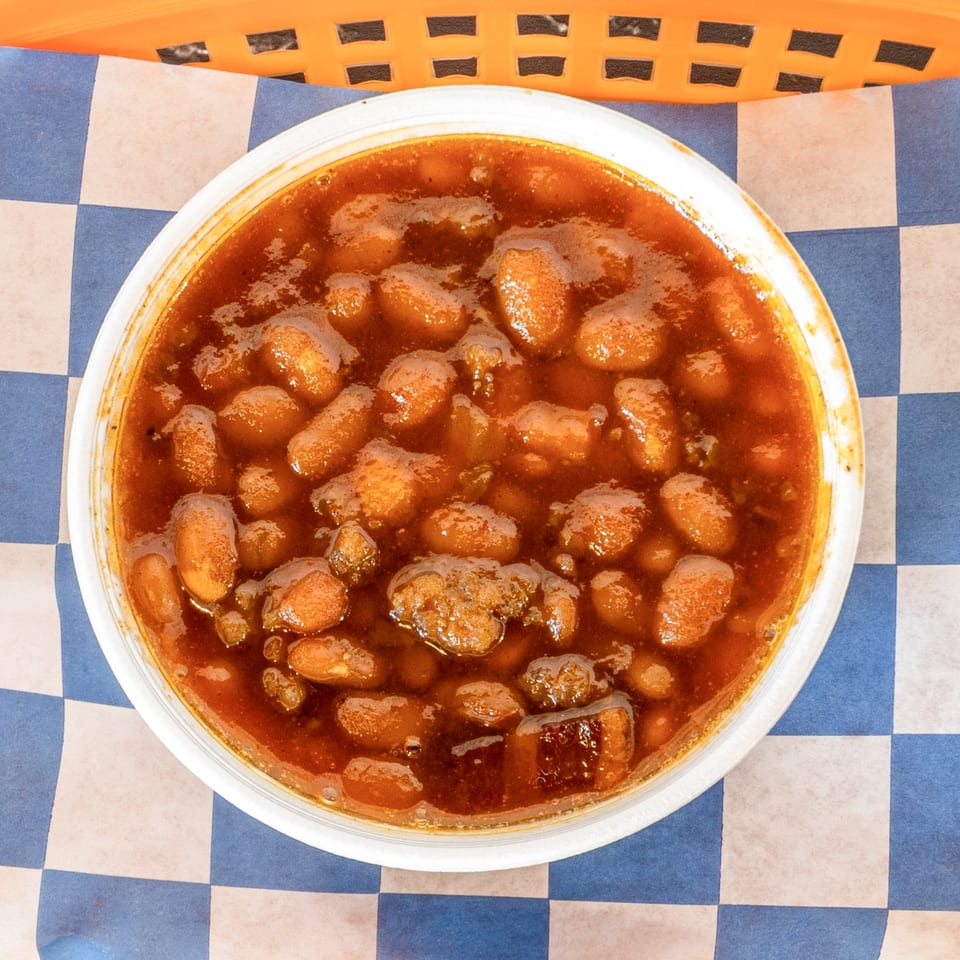 A white bowl of Suthern Cluck's baked beans with a rich, reddish-brown sauce on checkered paper.