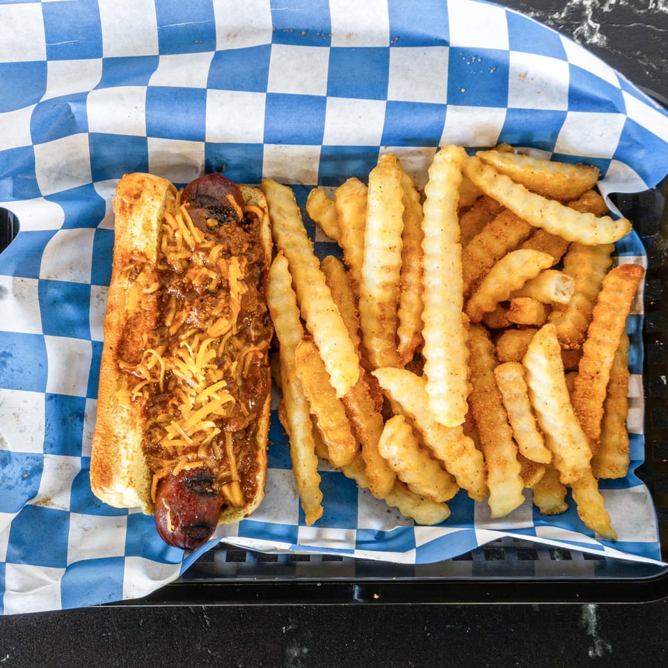 Chili cheese hot dog with crinkle-cut fries in a basket lined with blue and white checkered paper.