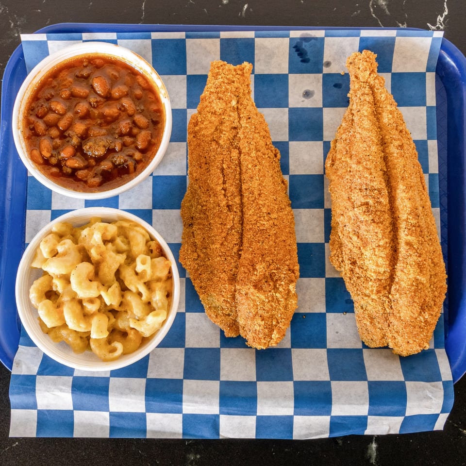 Two fried fish fillets, baked beans, and macaroni and cheese on a blue checkered tray.