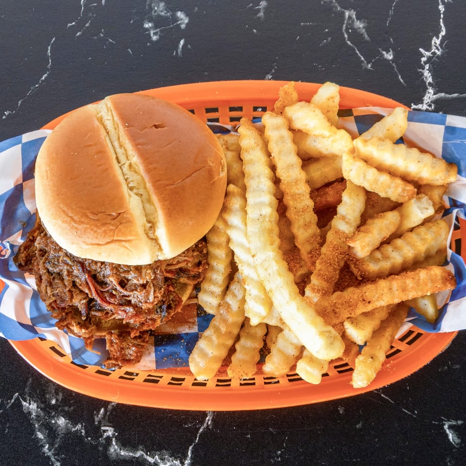 Pulled pork sandwich on a bun with a side of seasoned crinkle-cut fries in an orange basket.